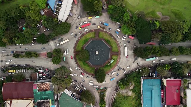 Top-down aerial slow-motion over Intramuros circular intersection with Simon de Anda Monument fountain, capturing vehicles navigating the roundabout surrounded by green lawns and residential areas.