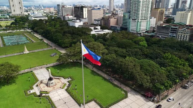 Slow motion aerial view of the Philippine flag waving proudly at Rizal Park in Manila. Urban skyline with high-rise buildings and green spaces frame this patriotic symbol in the heart of the capital.
