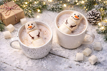 Two festive mugs of hot chocolate with marshmallow snowmen on a snowy winter table with holiday decorations