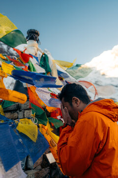 Himalaya, Nepal - 20 November 2025: View of a man in an orange jacket praying amidst vibrant prayer flags fluttering against the backdrop of snow-capped peaks.