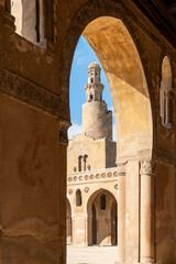 Ibn Tulun Mosque in Cairo viewed through arches. The mosque features a tall minaret and intricate...