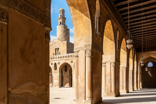 Ibn Tulun Mosque in Cairo viewed through arches. The mosque features a tall minaret and intricate architectural details. Blue sky visible in the background.
