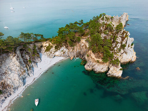 Aerial view of craggy cliffs meet the turquoise sea, a natural arch framed by verdant trees, creating a dramatic contrast of textures and tones, Crozon, Finistre, France.