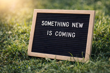 A black sign with white letters on grass announcing something new is coming