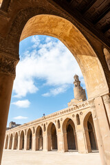 Ibn Tulun Mosque in Cairo viewed through arches. The mosque features a tall minaret and intricate...