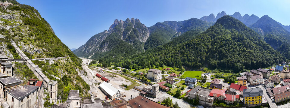 Aerial view of rugged mountains meet a quaint town with red-roofed buildings and an old mining complex, Cave del Predil, Friuli-Venezia Giulia, Italy.