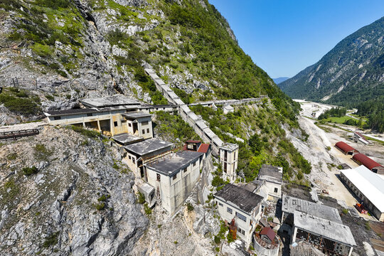 Aerial view of weathered buildings nestled against a steep, rocky mountainside, contrasted by the lush greenery of the surrounding valley, Rabelj, Friuli-Venezia Giulia, Italy.