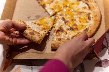 Hands reach for cheese pizza in a cardboard box on a table during dinner time