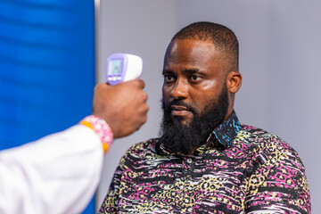 African male doctor uses a non-contact thermometer to check a male patient's temperature during a consultation in a hospital