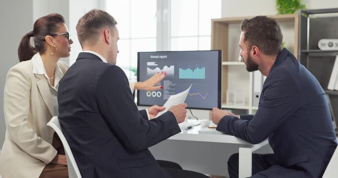 Business team meeting in office with report. Colleagues review data on a computer, studying charts for analysis. Team focuses on results during discussion. Solid team cooperation concept shown.