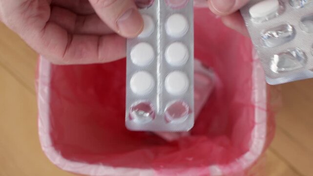 Closeup hands drop unused blister pack pills into red trash bin during daily medicine cabinet declutter and safety check.
