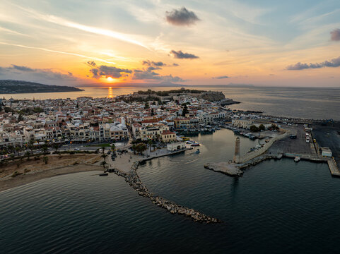Aerial view of the Venetian harbor embraced by the golden sunset and the old lighthouse, with the Fortezza fortress silhouetted against the horizon, Rethimno, Rethymno, Greece.