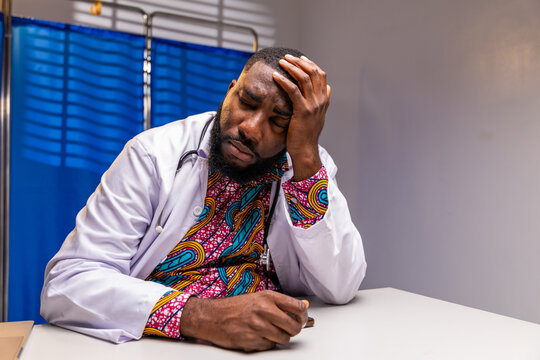 Stressed African male doctor with eyes closed, hands on temples sitting at his desk in a hospital, reflecting burnout and exhaustion in the profession