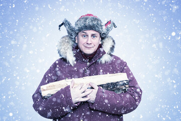 Funny man in a fur ushanka holding firewood during heavy snowfall. Winter survival concept, home heating in cold weather, rural lifestyle, humor, seasonal energy and warmth.