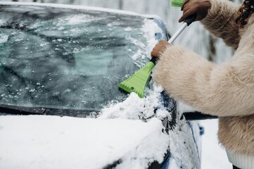 Woman using a green ice scraper to remove snow from the windshield of a car parked in a snowy environment during winter weather conditions