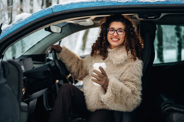 Young woman with curly hair wearing a fluffy coat is sitting in a car, holding a coffee cup, with snow-covered trees visible outside the window during winter