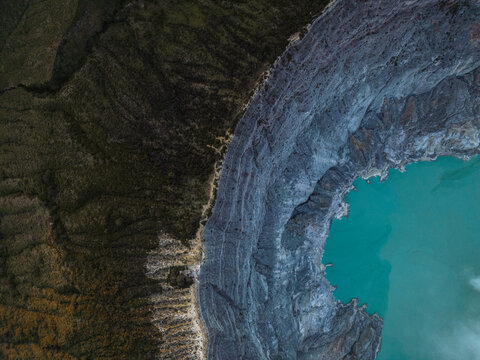 Aerial view of the stark, rugged terrain meeting the turquoise waters of the volcanic crater lake, creating a mesmerizing contrast of color and texture, Banyuwangi, East Java, Indonesia.