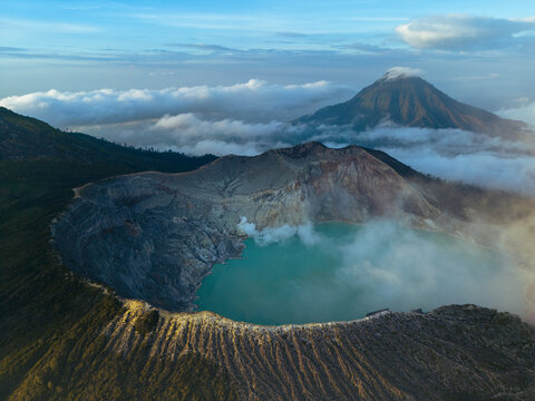 Aerial view of the majestic Ijen volcano crater lake, a mesmerizing turquoise jewel amidst the rugged, smoky terrain and distant peaks, Banyuwangi, East Java, Indonesia.