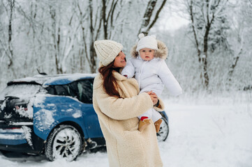 Obraz premium Woman in cozy winter coat holds child in snowy landscape near blue car, showcasing family bonding and joy during winter activities in a serene environment
