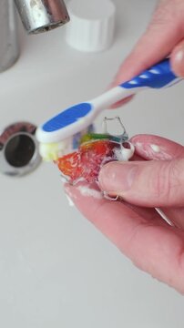Hand scrubs colorful orthodontic retainer with toothbrush and toothpaste in sink, demonstrating daily oral appliance care.