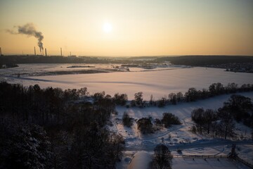 Sweeping aerial vista of a frozen winter realm. Golden hour sunlight bathes the vast snowy plain, creating a stark contrast with distant industrial silhouettes against the sky