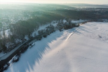Aerial view of a frozen river winding through a snowy landscape under the soft morning sun. Bare trees cast long shadows across the pristine snowfields, hinting at a peaceful winter dawn.