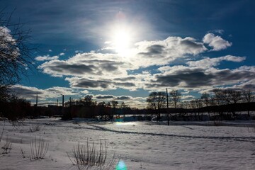 Sunlight pierces through dramatic clouds, illuminating a vast snow-covered landscape with scattered trees and distant industrial structures, a true winter's stark beauty