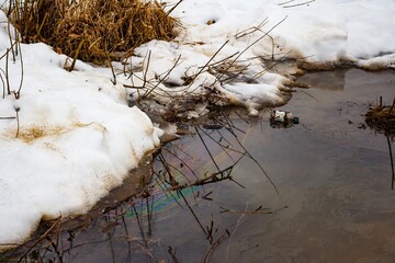 Iridescent oil film pollutes a murky winter river, reflecting a stark warning of ecological damage. Dirty snow and debris highlight environmental neglect