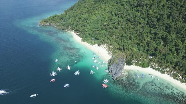 Aerial drone footage showcasing Seven Commandos Beach from a different perspective, featuring white sand, clear turquoise water, tourist boats and dramatic limestone cliffs in El Nido, Philippines.