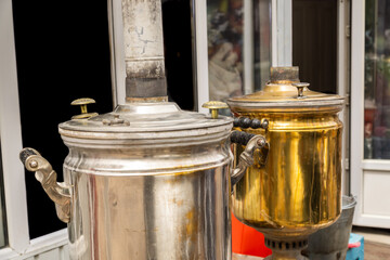 Two samovars, one silver and one brass, sit outdoors near a charming building. Nostalgic atmosphere evokes traditional tea culture.