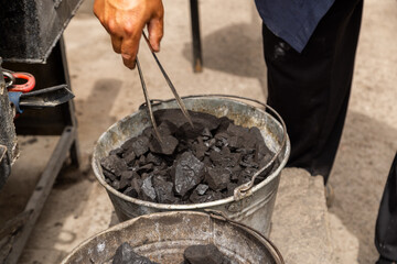 Focused man diligently uses tongs to pick up coal in outdoor workshop. Purposeful action reflects rustic work environment and industrious mood.