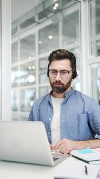 Corporate operator in headset working in customer support service on helpline telesale talking using laptop in business office. Agent of call center advises client answering questions. Vertical video