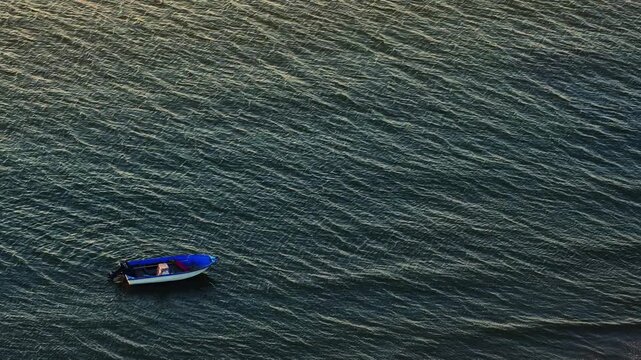 Small blue motorboat on rippled water and ocean sea waves at sunset with golden sunlight reflection creating calm peaceful solitary mood