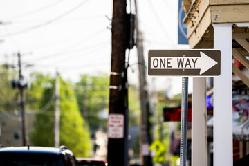 White rectangular one way street sign with black arrow indicating traffic direction mounted on pole at urban intersection with buildings and trees in background. 