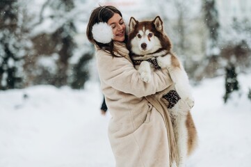 Young woman with long dark hair wearing a beige coat and earmuffs is joyfully holding a fluffy...