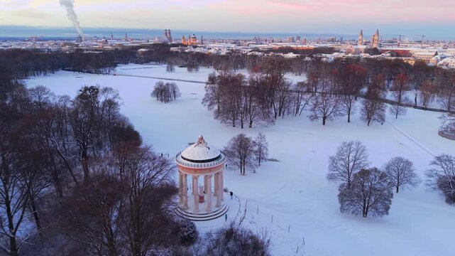 Luftaufnahme vom winterlichen Muenchen. Blick vom verschneiten Englischen Garten mit Monopteros auf die historische Altstadt und das Stadtzentrum bei einem wunderschoenen Sonnenaufgang