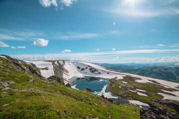 Scenic sunlit landscape with alpine lake in rocky snowy cirque near stone hill top in sunny day during thaw. Ice floats in mountain lake among rocks with view to forest mountain range under bright sun