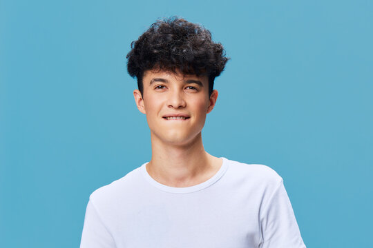 young man with curly hair biting lip posing confidently in white t-shirt against blue background, casual style, playful expression, studio portrait.