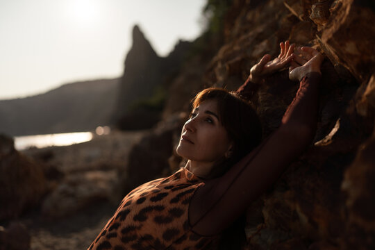 Woman nature sunset. Beautiful young woman relaxing by rocky shore looking up at sky during warm golden hour. Peaceful reflective mood. - Powered by Adobe
