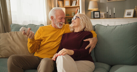 A senior couple sits on a couch, smiling and laughing while watching TV. They hold a remote control...