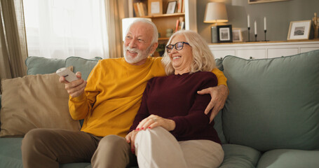 Obraz premium A senior couple sits close together on a couch watching television. They share smiles and seem to enjoy their time together. The living room has warm lighting and a cozy feel.