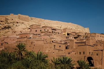Ancient adobe fortress village in arid desert landscape with palm trees, traditional earthen architecture and mountain backdrop under clear blue sky.