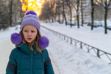 Girl wears knit hat with pompom and puffy jacket on snowy street with trees and fence. Bright sky and blurred buildings are visible in distance.