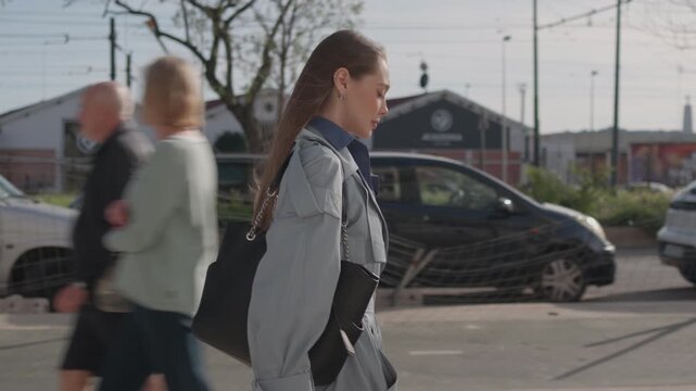 Young businesswoman in blue coat walking with purse along city sidewalk with confident expression. Trees and pedestrians are visible on sunny summer day