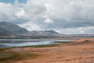 A peaceful landscape with Lake Karakol and mountains under a cloudy sky. In the foreground there is dry grass and a path. Natural beauty, nature, spaciousness, serenity in the East Kazakhstan region
