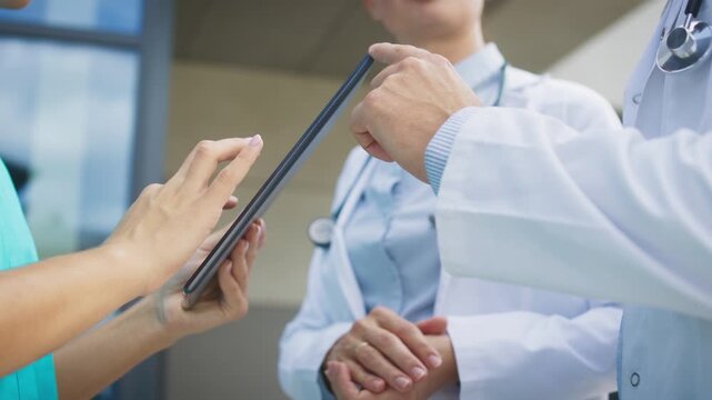 Side view showing tablet device. Female nurse tapping on screen with fingers. Two doctors discussing treatment plan calmly in front. Standing outside large hospital area. Modern technology concept.
