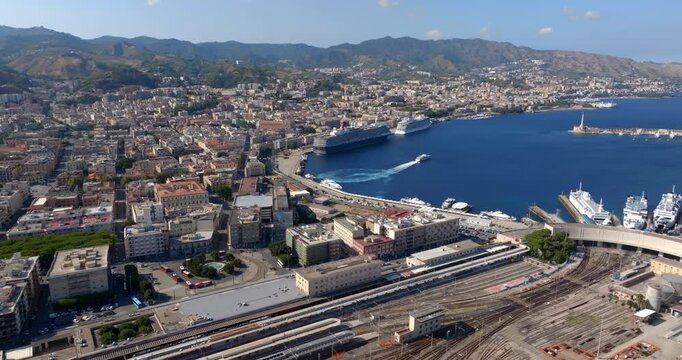 Aerial view of the city port of Messina, Sicily, Italy. Panorama of a crescent-shaped harbour. Large cruise ships and various vessels are docked along the quay. The train station is in foreground.