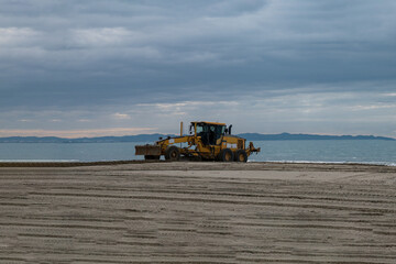 Fototapeta premium Bulldozer leveling sand on coastal beach near the sea under dramatic cloudy sky, construction machinery, coastal maintenance and environmental management concept.