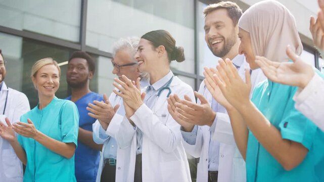 Smiling medical staff clapping hands while standing outside clinic under bright daylight. Confident group celebrating progress and sharing uplifting energy. Professionals enjoying supportive moment.