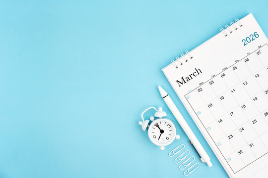 March 2026, Monthly desk calendar for 2026 year and alarm clock with paper clips on blue color background.